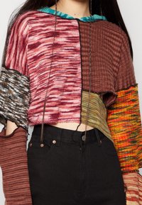 Sweter o skróconym kroju robiony na drutach z wzorem patchworkowym w odcieniach różu, brązu i zieleni, charakteryzujący się różnymi teksturami i otwartymi rękawami. W zestawie z czarnymi spodniami.