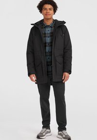 Ung man i svart huva parkas, blå och svart rutiga skjorta, svarta byxor och grå sneakers, som står mot en vit bakgrund.