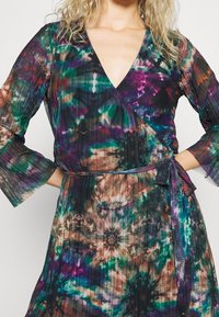 Robe portefeuille plissée multicolore avec un col en V et de longues manches évasées. Elle possède une ceinture à nouer et des motifs abstraits vibrants en vert, violet et marron.