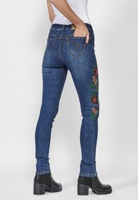 Skinny jeans in dark blue denim, featuring colorful floral embroidery along the left leg, designed with five pockets and sturdy stitching.