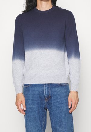 Personne portant un pull à manches longues à col rond avec un dégradé bleu marine à gris clair, associé à un jean bleu, debout devant un fond uni.
