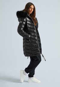 Melnā pūkainā mēteli ar kažokādas apdari ap kapuci, quilted dizains, garš piegriezums un priekšējā rāvējslēdzēju aizdare. Apvienots ar melnajām sporta biksēm un baltām sporta apaviem.