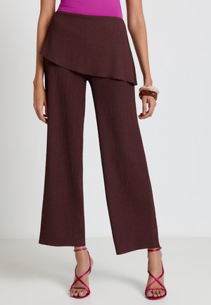 Frau trägt braune, strukturierte, weit geschnittene Hosen mit asymmetrischem Rocküberwurf, kombiniert mit pinken Riemchenabsätzen und Armbändern.