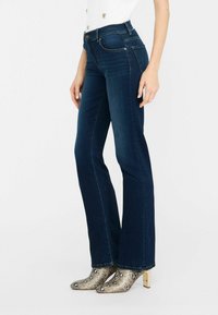 Jeans acampanados de denim azul oscuro con bolsillos delanteros y traseros, con costuras doradas y cierre de botón, combinados con botas de tobillo de estampado de serpiente metálico.