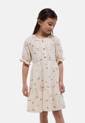 Jeune fille regardant vers le bas, portant une robe crème avec des boutons, des manches bouffantes et un motif floral et d'abeille sur un fond uni.