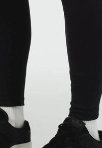 Leggings neri con una texture liscia, abbinati a sneakers nere con suola bianca e branding in evidenza. Calzini bianchi visibili sopra le sneakers.