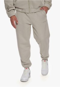 Beige sweatpants with elastic cuffs and a drawstring waist, featuring discreet logo embroidery on the front and worn with white sneakers.
