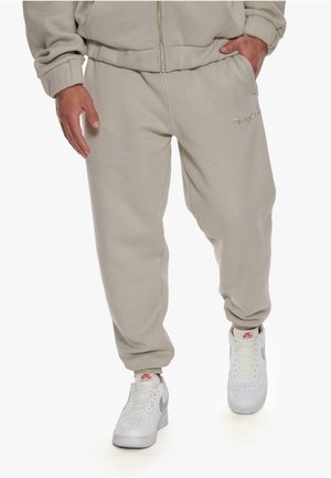 Beige sweatpants with elastic cuffs and a drawstring waist, featuring discreet logo embroidery on the front and worn with white sneakers.