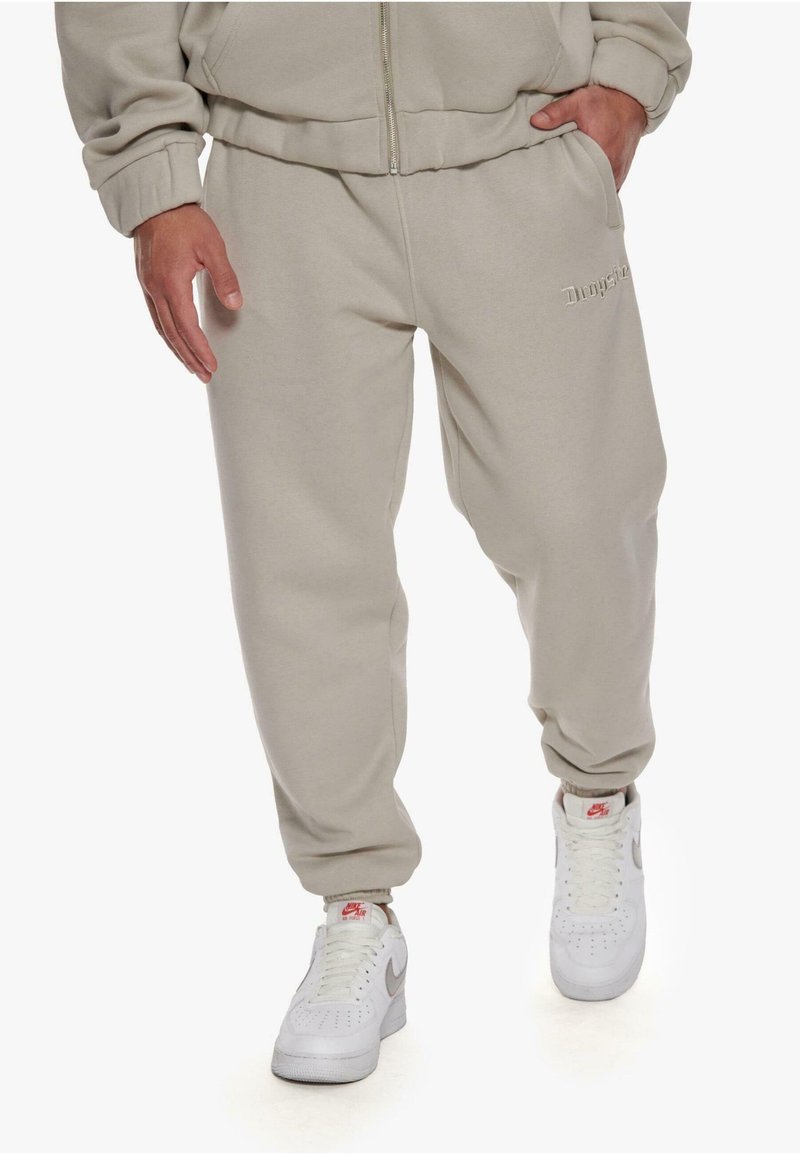 Beige sweatpants with elastic cuffs and a drawstring waist, featuring discreet logo embroidery on the front and worn with white sneakers.