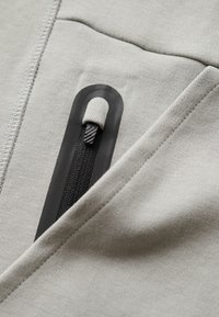 Detalle de bolsillo con cremallera en gris claro sobre una tela suave, con una cremallera negra que presenta un tirador texturizado y costuras lisas.