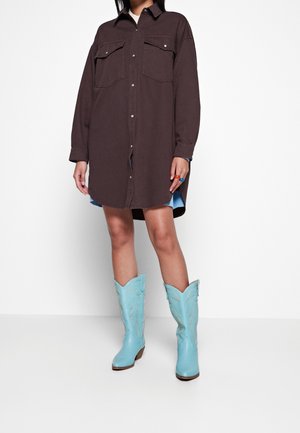 Femme portant une robe chemise marron foncé boutonnée avec deux poches poitrine et des bottes de cowboy bleu clair montant jusqu'aux genoux, debout devant un fond blanc.