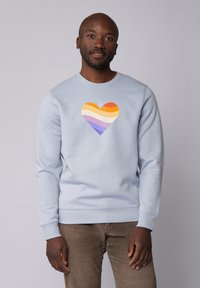 Lichtblauwe sweatshirt met een hartafbeelding met horizontale strepen in oranje, geel, crème, paars en lavendel. Gladde textuur.