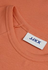 Camiseta de algodón acanalada en coral tenue con cuello redondo y una etiqueta blanca con texto azul. La textura parece suave y lisa.