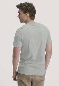 Jonge man met golvend haar, gekleed in een lichtgrijze T-shirt met korte mouwen en beige corduroy broeken, die iets naar links is gedraaid.