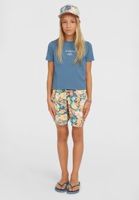 Blaues T-Shirt mit weißem Logo, kurze Hosen mit Blumenmuster in Orange, Gelb und Grün sowie blaue Flip-Flops. Die Cap hat ein buntes Blumenmuster.