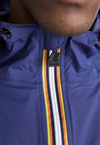 K-Way jacket with a dark blue nylon exterior, featuring a zipper with vertical stripes in orange, yellow, and navy. Includes an adjustable hood.