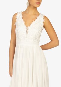 Weißes Kleid mit tiefem Ausschnitt, Spitze an den Schultern und einem fließenden, plissierten Rock. Enthält transparente Einsätze und ist bodenlang.