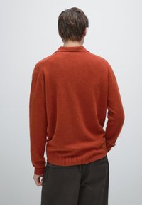 Sweter w kolorze rdzy o miękkim, teksturowanym wykończeniu, z kołnierzykiem polo oraz ściągaczami na rękawach i u dołu, noszony na ciemnych spodniach.