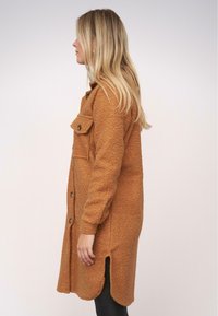 Barna gyapjúkabát texturált felülettel, elülső gombos záródással, mellzsebekkel és oldalsó hasítékokkal, fekete leggings fölött viselve.