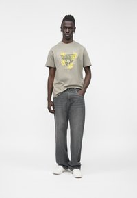 Pelēka kokvilnas T-krekls ar ziedu grafiku un uzrakstu "GUESS", kopā ar vaļīgām tumši pelēkām džinsu biksēm un baltām sporta kurpēm. Vienkāršs fons.