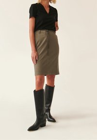 Haut noir à manches courtes, jupe verte olive jusqu'aux genoux avec cordon de serrage, et bottes en cuir noires jusqu'aux genoux avec bouts pointus et talons bas.