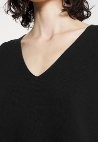 Zwarte gebreide trui met een diepe V-hals en geribbelde textuur. Focus op de nek- en schouderpartij, met nadruk op de zachtheid van het materiaal.