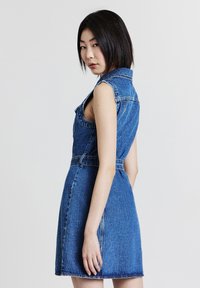 Robe en denim, sans manches avec col, de couleur bleu clair, corsage ajusté, jupe évasée, détails de couture visibles et poches latérales.