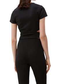 Černý žebrovaný crop top s krátkými rukávy a výřezem na zádech, kombinovaný s úzkými černými kalhotami. Hladká textura, obepínající střih.