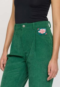 Pantalon en velours côtelé vert à taille haute, avec un plis à l'avant et une étiquette colorée cousue sur la poche. Tissu texturé et résistant.