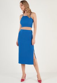 Kétrészes szett élénk kék anyagból, amely egy testhezálló crop topból és egy karcsú midi szoknyából áll, oldalsó résvágással. A modellen rózsaszín magassarkú cipő van.