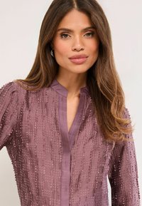 Lila texturált blúz finom mintázattal, mandzsettagallérral és egyszerű V-nyakkivágással. Puha anyag, változatos felületi részletekkel.