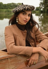 Ziloņu raksta berete, brūna zamšādas jaka ar pogu slēgšanu, ribota tumši brūna džemperis, zeltainas masīvas auskari un gredzeni uz rokām.