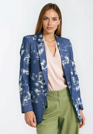 Niebieska marynarka z białymi abstrakcyjnymi wzorami, zapinana na jeden guzik, z klapami z wycięciami, zestawiona z jasnoróżową bluzką i oliwkowozielonymi spodniami.