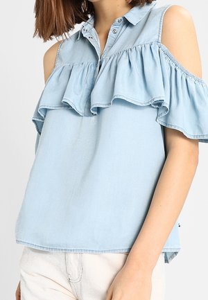 Vrouw draagt lichtblauwe cold-shoulder blouse met ruchtdetails en knopen bij de kraag, gecombineerd met lichtgekleurde broek.
