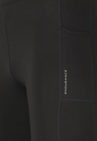 Svarta atletiska leggings av elastiskt material, med en sidoficka och texten "ENDURANCE" i vitt. Enkelt och stilrent design med platta sömmar.