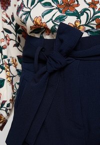Pantalon bleu marine taille haute avec un tissu texturé et un détail de nœud à l'avant de la taille. Chemisier à motif floral avec manches longues visibles.