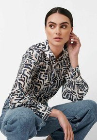 Frau mit dunklem Haar in einem gemusterten Langarmshirt und blauer Jeans sitzt mit einer Hand nahe ihrem Gesicht und blickt nach links vor einem weißen Hintergrund.