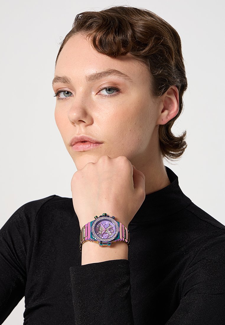 Montre colorée avec un cadran violet, un bracelet métallisé multicolore, un visage rond et des marquages détaillés, portée sur une main.