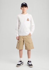 White long-sleeve shirt with orange graphic, tan cargo shorts, and black sneakers with white accents. The cap is navy with a logo.