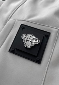 Grijze stoffen oppervlakte met een getextureerde zwarte vierkante patch met een zilvergegoten leeuwenkop-insigne en vier zilveren klinknagels in de hoeken.