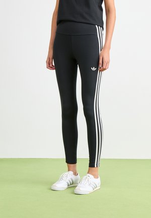 Cilvēks, kurš valkā melnas Adidas legingi ar baltām trim svītrām sānos un baltas Adidas kedas, stāv uz zaļas grīdas.