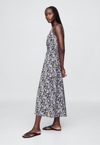 Maxikleid mit Blumenmuster in Schwarz, Weiß, Lila und Grün. Verfügt über dünne Träger und ein fließendes Design. Kombiniert mit schwarzen Flip-Flops.