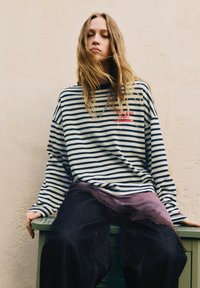 Jong persoon met lang blond haar draagt een zwart-wit gestreept lang shirt met lange mouwen en donkere broek, zittend tegen een beige, gestructureerde muur.