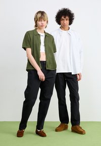 Two individuals pose in casual attire. One wears a green short-sleeve button-up over a white crop top, black pants, and brown shoes. The other has a white striped shirt, black pants, and brown shoes.