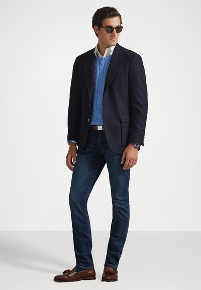 Ναυτικό σακάκι, ανοιχτό μπλε πουλόβερ με πλέξη καμπάνας, λευκό πουκάμισο με γιακά, σκούρο τζιν, καφέ loafers και στρογγυλά γυαλιά ηλίου. Κλασικό, κομμένο εφαρμοστό.