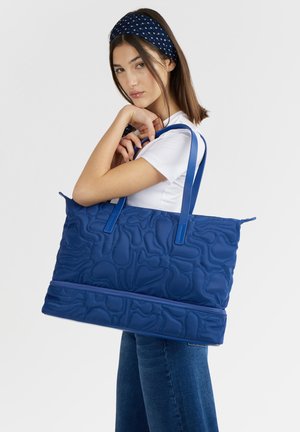 Giovane donna con camicia bianca e fascia per capelli che porta una grande borsa trapuntata blu con doppi manici e tasca con cerniera.