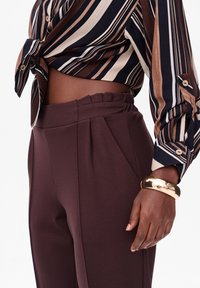 Chemise rayée avec des couleurs marron et marine, nouée à la taille ; pantalon plissé marron ; bracelet jonc en or ; textures de tissu lisses.