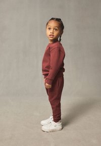 Sweat-shirt rouge en coton et pantalon assorti, avec des poignets côtelés et une coupe décontractée. Porté avec des chaussures de sport blanches. Enfant debout de profil.