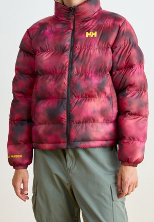 Modelo con una chaqueta acolchada Helly Hansen estampada en rojo y negro, con cuello alto y cremallera negra, combinada con pantalones cargo de color verde oliva.