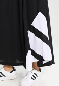 Svart kjol med vita diagonala ränder i nederkanten, tillverkad av lättviktsmaterial, i kombination med vita sneakers med svarta detaljer.